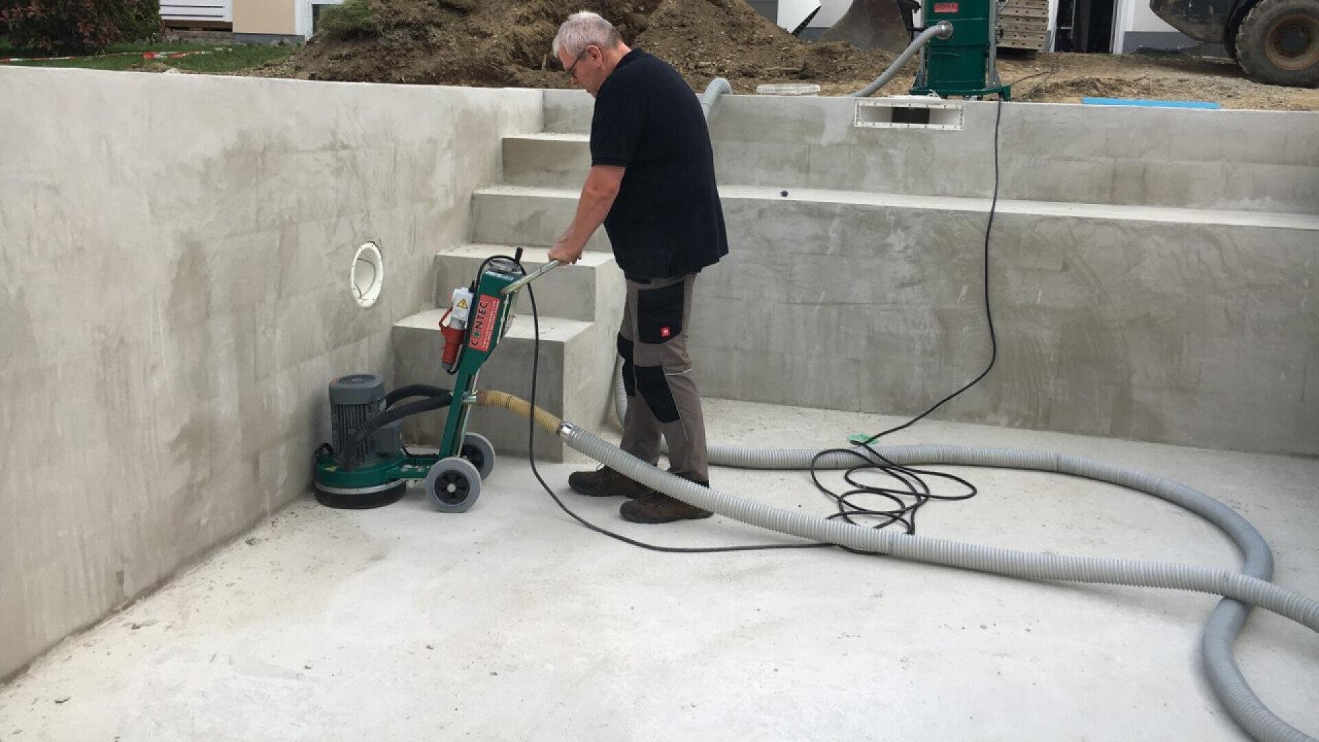 A person uses a floor grinding machine to smooth the concrete surface inside an empty swimming pool under construction. Hoses and cables are connected to the equipment.