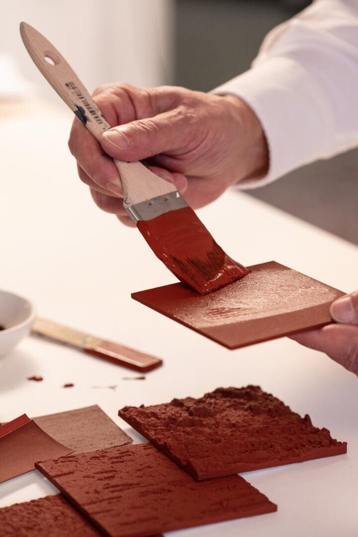 Eine Person trägt mit einem Pinsel rotbraune Farbe auf eine quadratische Fliese auf. Auf dem Tisch in der Nähe liegen mehrere strukturierte und glatte Fliesen.