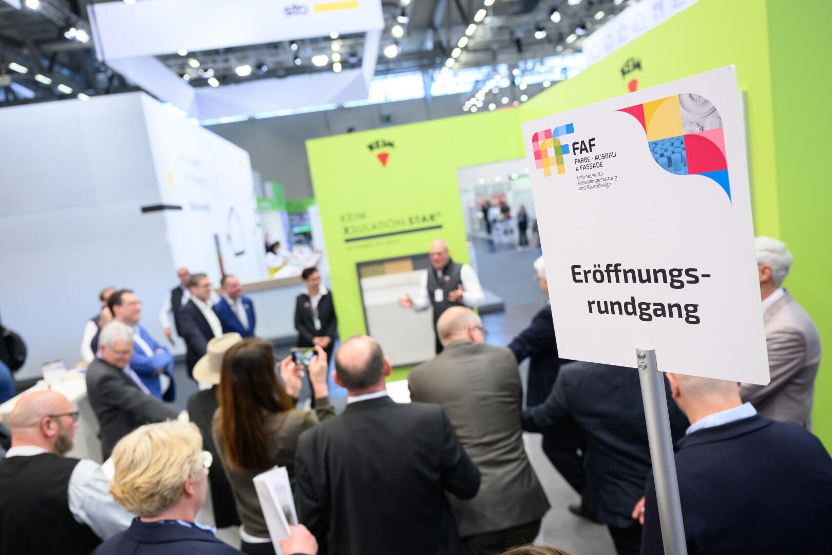 Eine Gruppe von Menschen versammelt sich um einen Redner bei einer Indoor-Ausstellung. Im Vordergrund steht ein Schild mit der Aufschrift „Eröffnungsrundgang“ auf Deutsch und auf dem Schild befinden sich bunte Grafiken.
