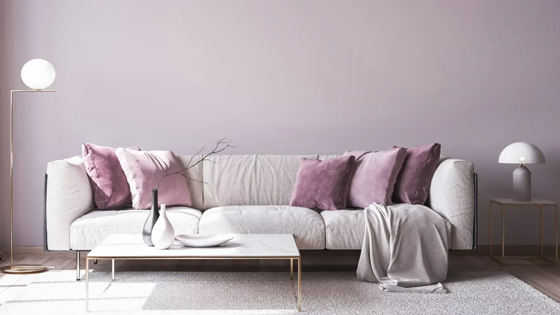 Ein modernes Wohnzimmer mit einem hellgrauen Sofa, dekoriert mit malvenfarbenen und rosa Kissen, einer Überwurfdecke, einem weißen Marmor-Couchtisch mit Vasen, einer Stehlampe, einem Beistelltisch mit einer Lampe und einer hellvioletten Wand.