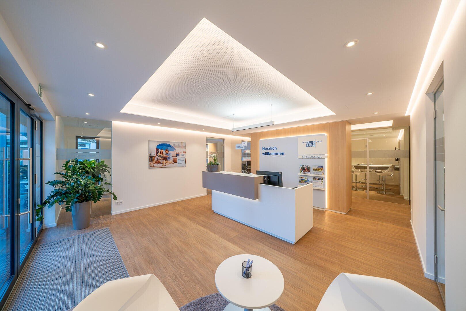 Ein moderner Büroempfangsbereich mit einem weißen Schreibtisch, einem Willkommensschild, hellem Holzboden, hellen Deckenleuchten, Zimmerpflanzen und einem kleinen Tisch mit zwei weißen Stühlen im Vordergrund.