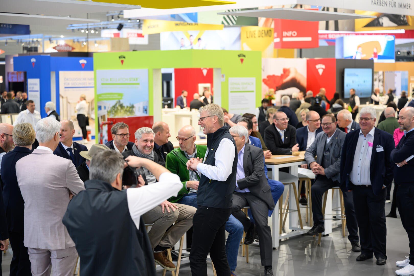 Eine Gruppe von Menschen, hauptsächlich Männer, versammelt sich auf einer lebhaften Veranstaltung oder Messe. Einige sitzen an Stehtischen, andere stehen und unterhalten sich. Bunte Stände und Informationsschilder prägen den Hintergrund.