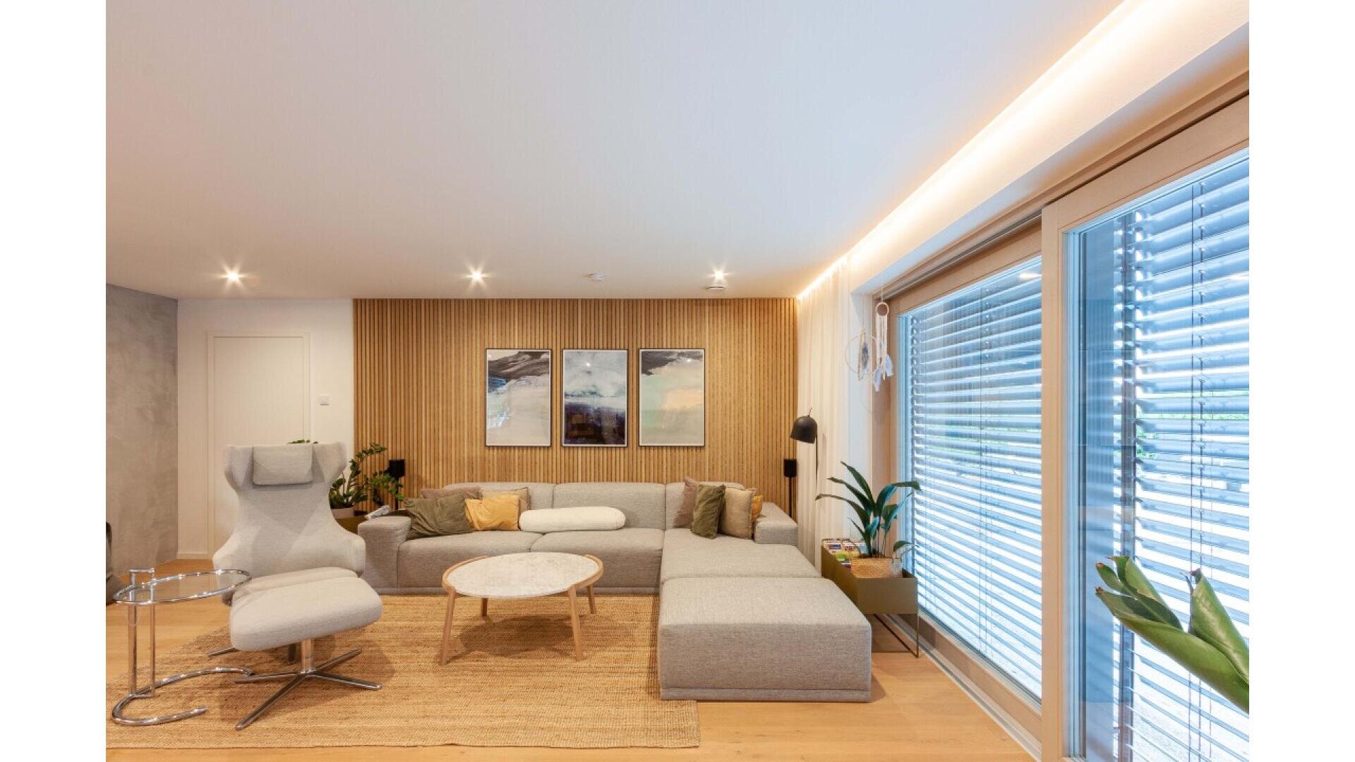 A modern living room with a gray sectional sofa, light armchair, round coffee table, wooden accents, two framed pictures on a wood-paneled wall, plants, and large windows with blinds letting in natural light.