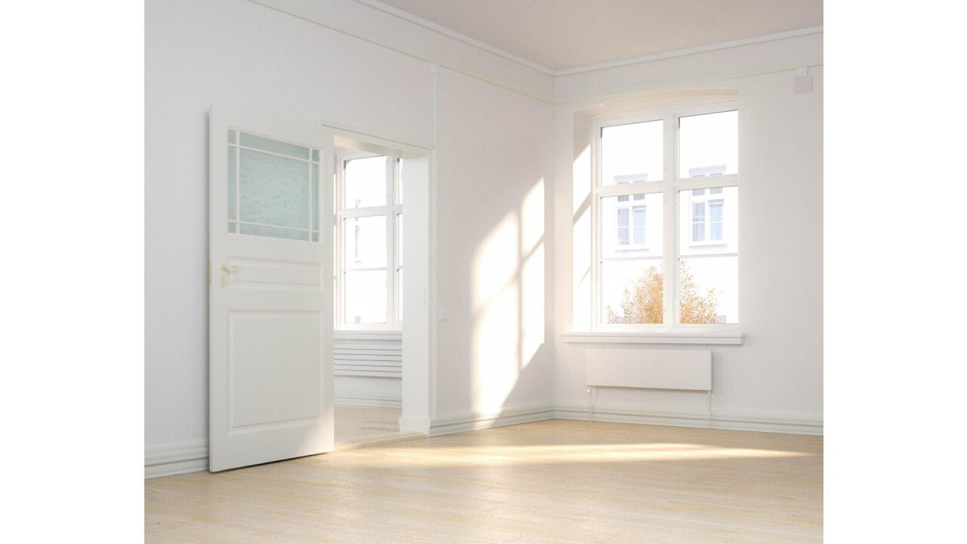 Bright, empty room with white walls, large windows letting in sunlight, light wooden floor, and a white door partially open. Outside the windows, some trees and building details are visible.