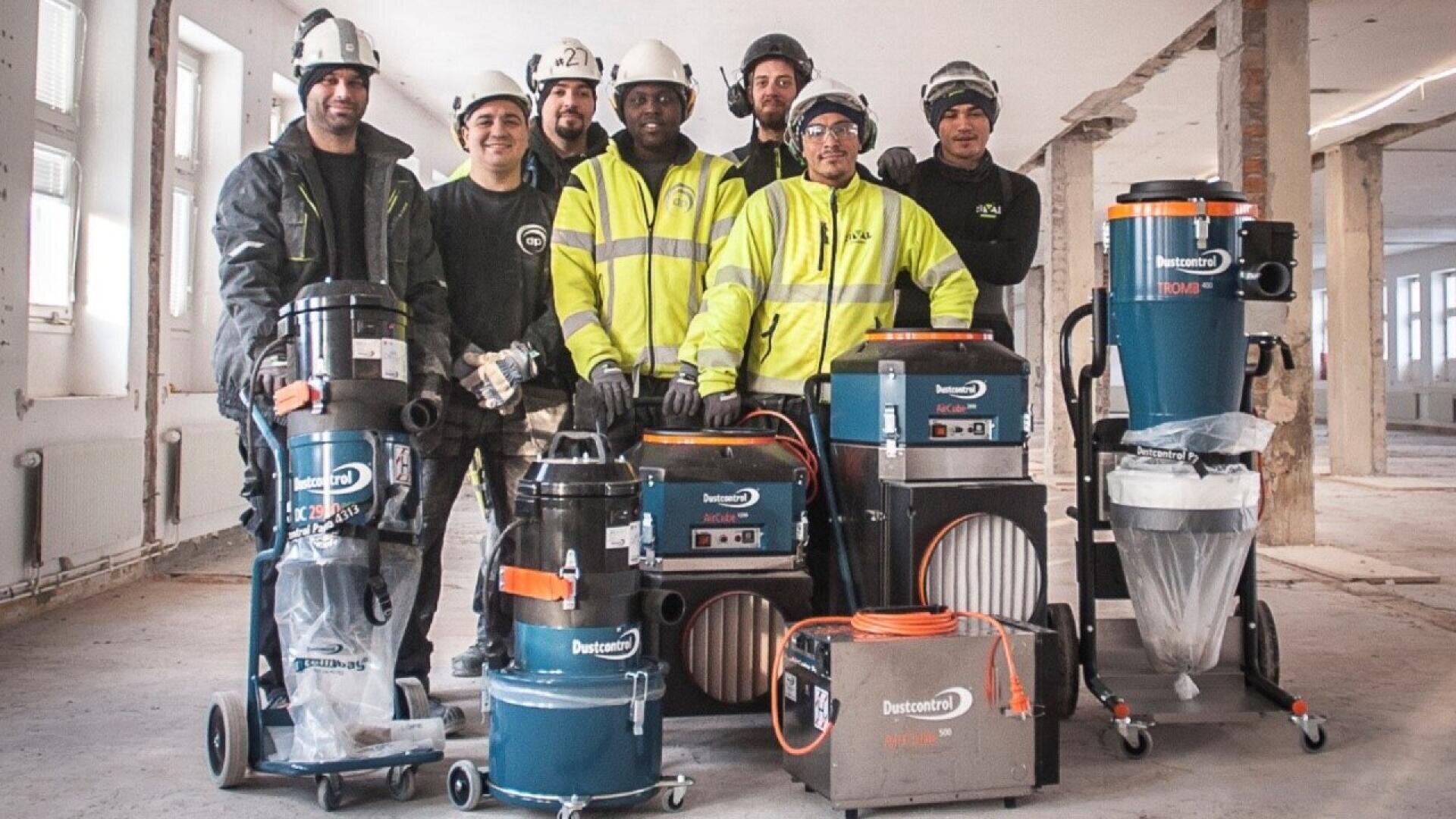 A group of seven construction workers in safety gear pose indoors with industrial vacuum machines and cleaning equipment in a partially renovated building.