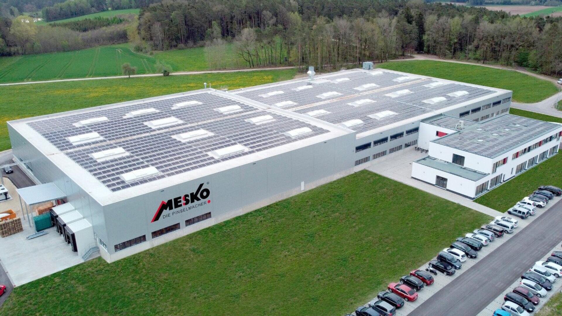 Aerial view of a large industrial building with solar panels on the roof, marked MESKO, surrounded by greenery, a parking lot with cars, and adjacent forested area.
