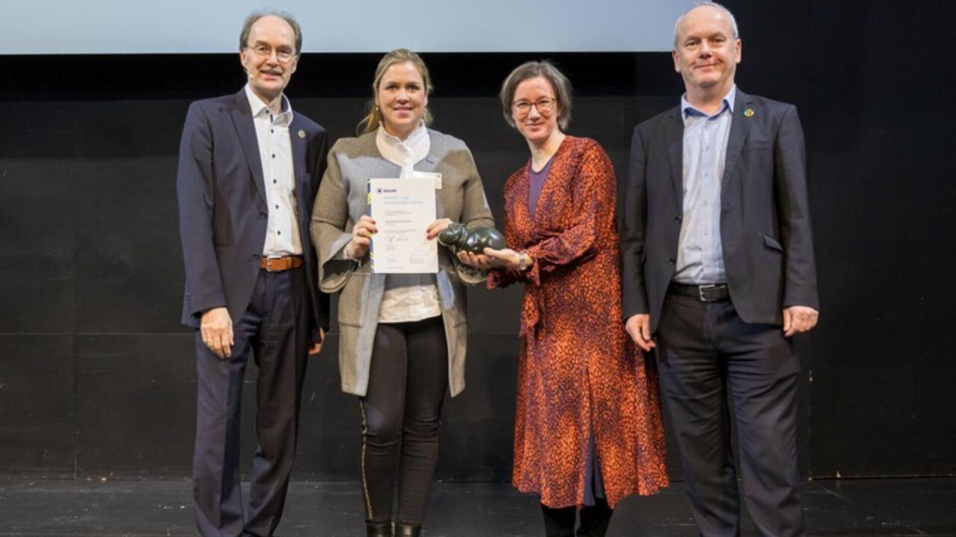 Four people stand on a stage; one woman holds a certificate, another holds a black object, while the two men on either side smile. They appear to be at an award or recognition event.