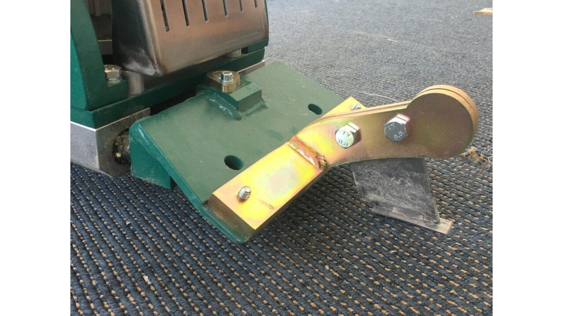 A close-up of a metal floor scraper tool attached to a green industrial machine, positioned on a dark blue carpeted surface. The scraper blade is angled downward, and bolts are visible on the metal attachment.