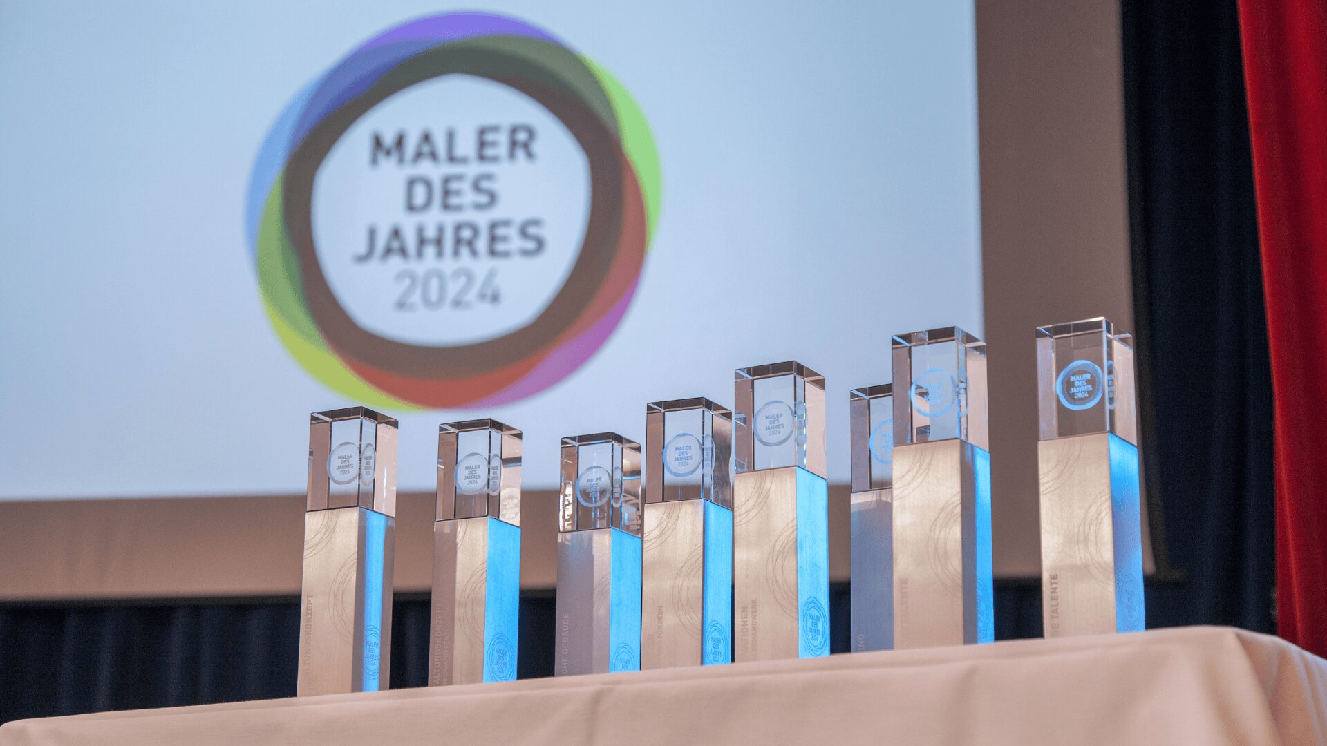 Bei einer Veranstaltung werden mehrere gläserne Auszeichnungen auf einem Tisch vor einem Bildschirm präsentiert, auf dem das Logo des Malers des Jahres 2024 zu sehen ist.