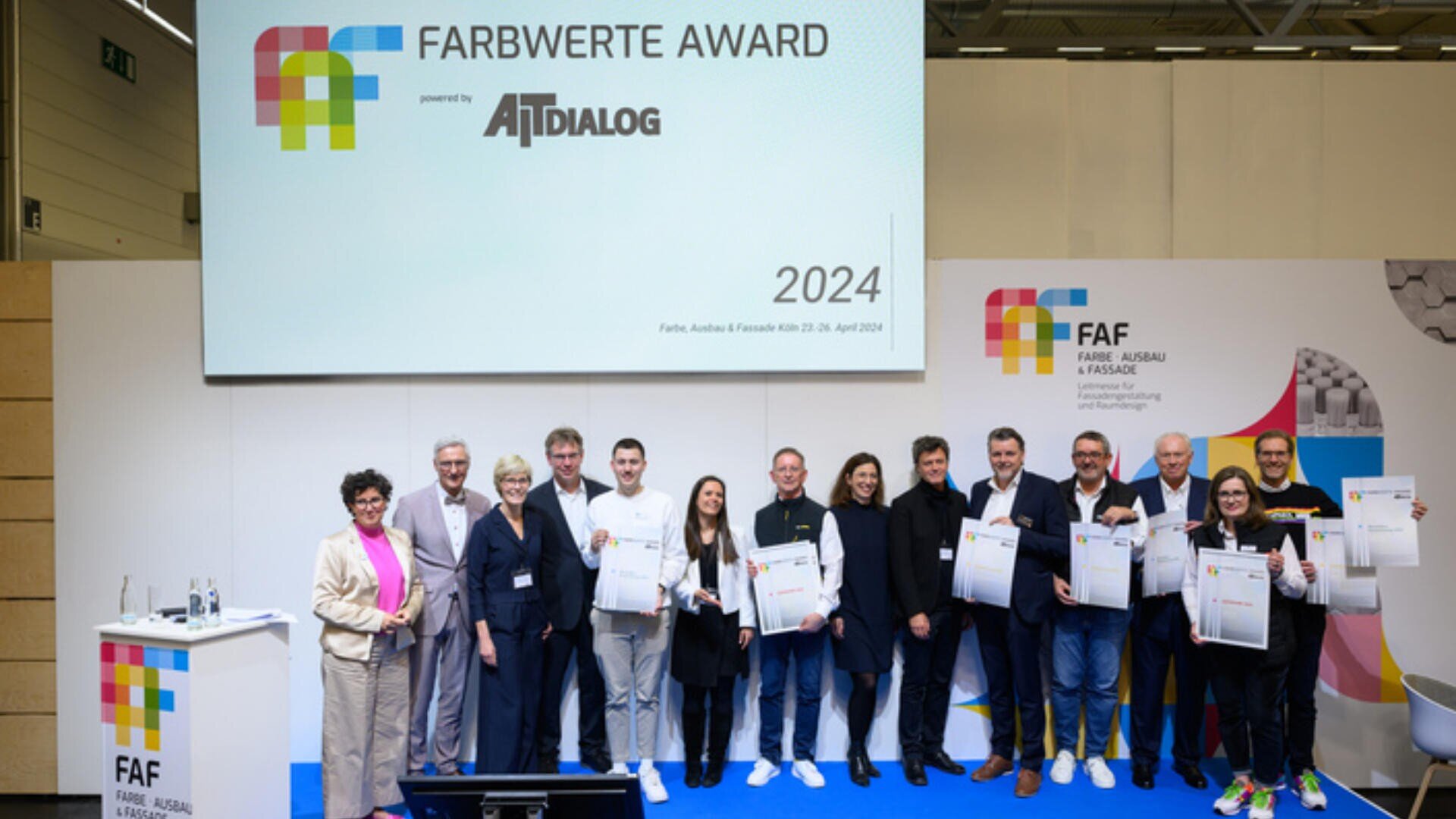 Bei der Verleihung des Farbwerte Awards 2024 steht eine Gruppe von Menschen mit Urkunden auf der Bühne, dahinter sind eine große Leinwand und bunte Logos zu sehen.