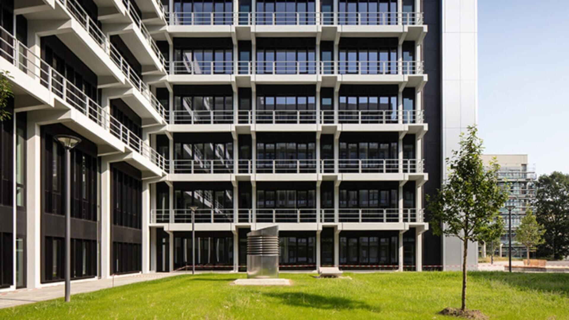 A modern building with black and white balconies overlooks a green lawn with a few small trees and a cylindrical metal sculpture in the center.