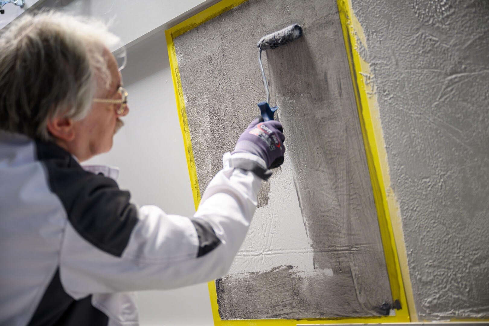 Eine Person mit Handschuhen und Schutzkleidung trägt mit einer Farbrolle graues Material auf eine strukturierte Oberfläche auf, die mit gelbem Klebeband umrahmt ist.