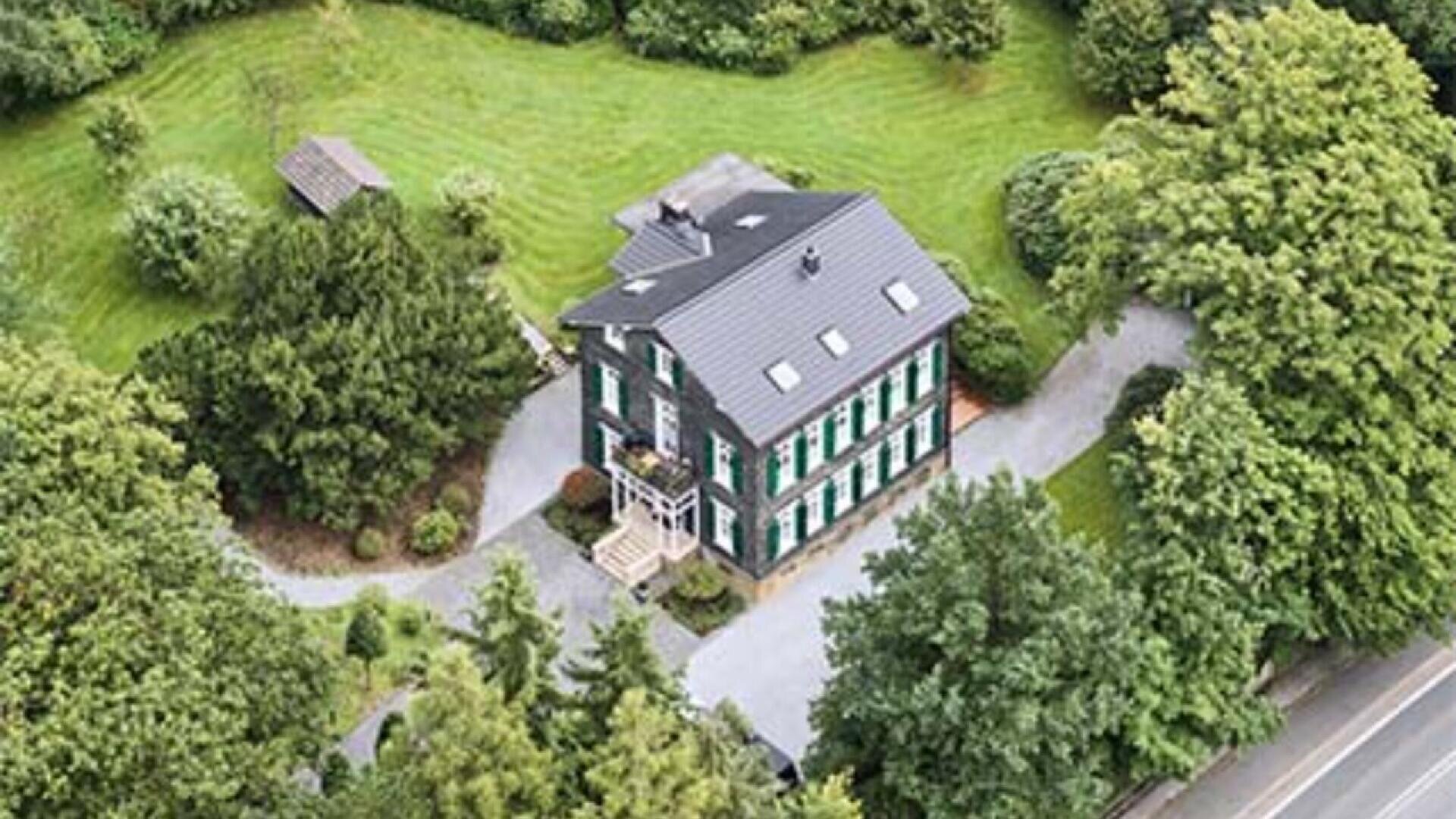 Aerial view of a large, dark-colored house with green shutters, surrounded by trees and a well-maintained lawn, with a driveway leading to a road on the right side.