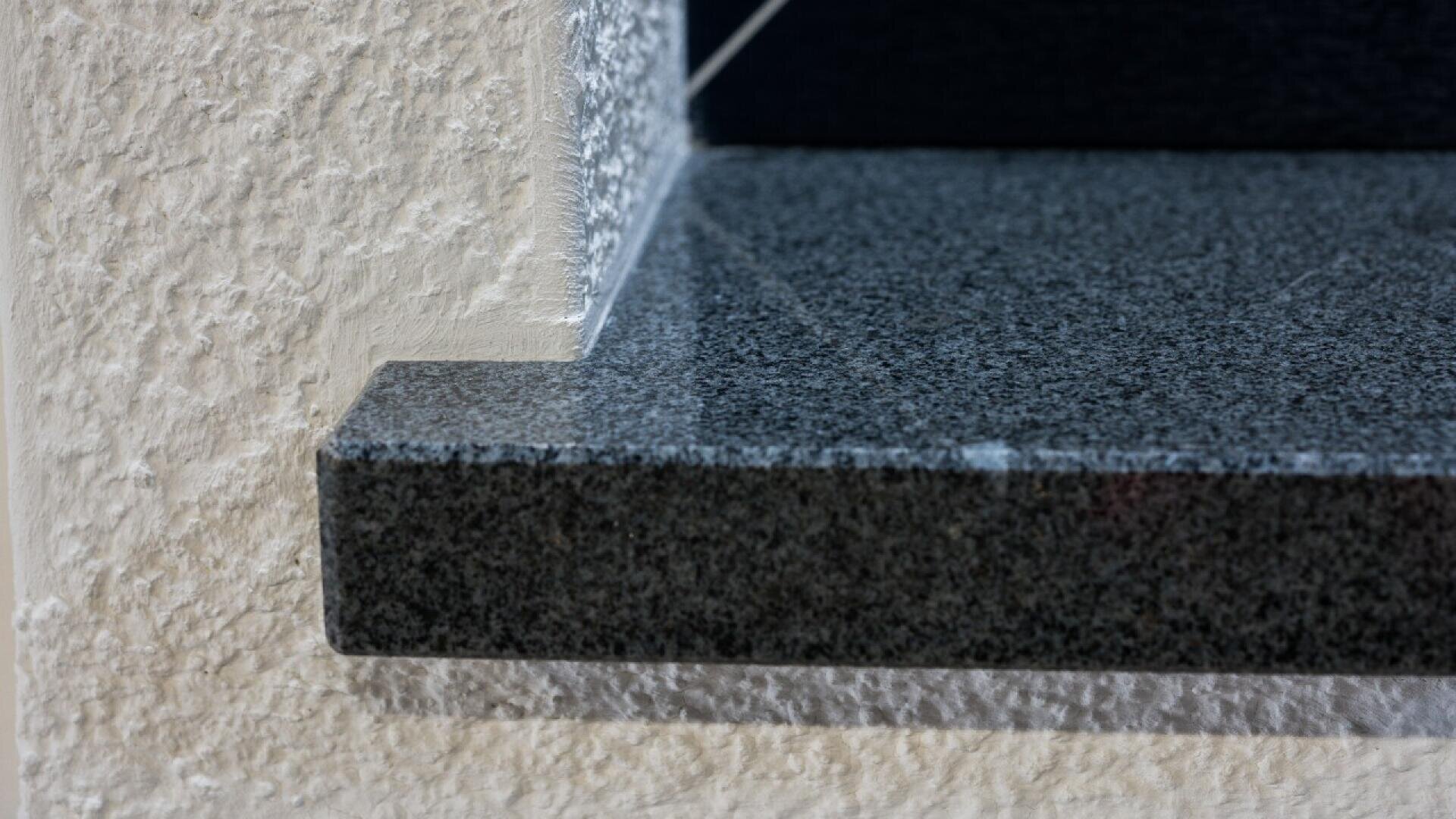 Close-up of a polished dark granite windowsill extending from a textured, off-white stucco wall, showcasing the contrast between smooth stone and rough wall surface.