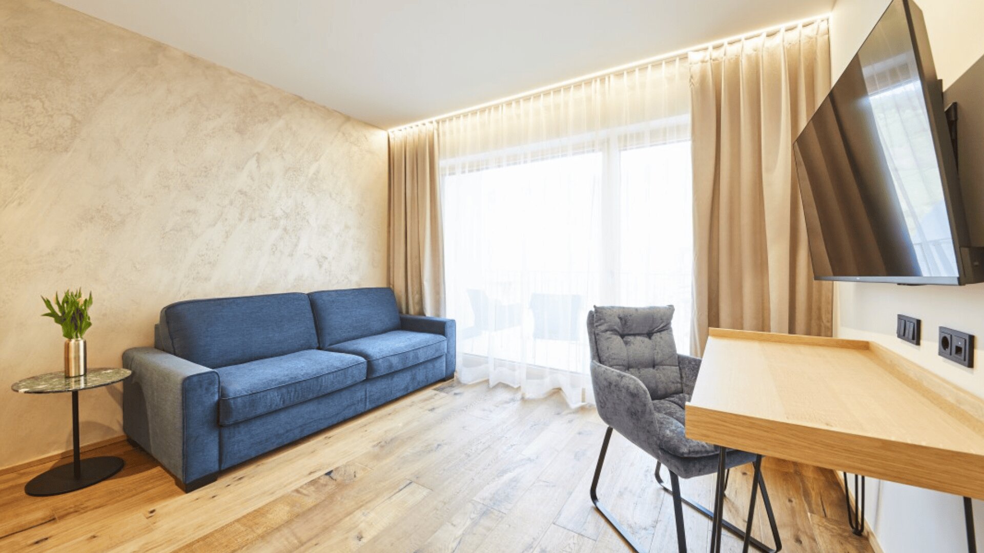 A modern, bright living room with a blue sofa, a small round glass table with flowers, a wooden desk with a gray chair, a large wall-mounted TV, wood flooring, and floor-to-ceiling windows with beige curtains.