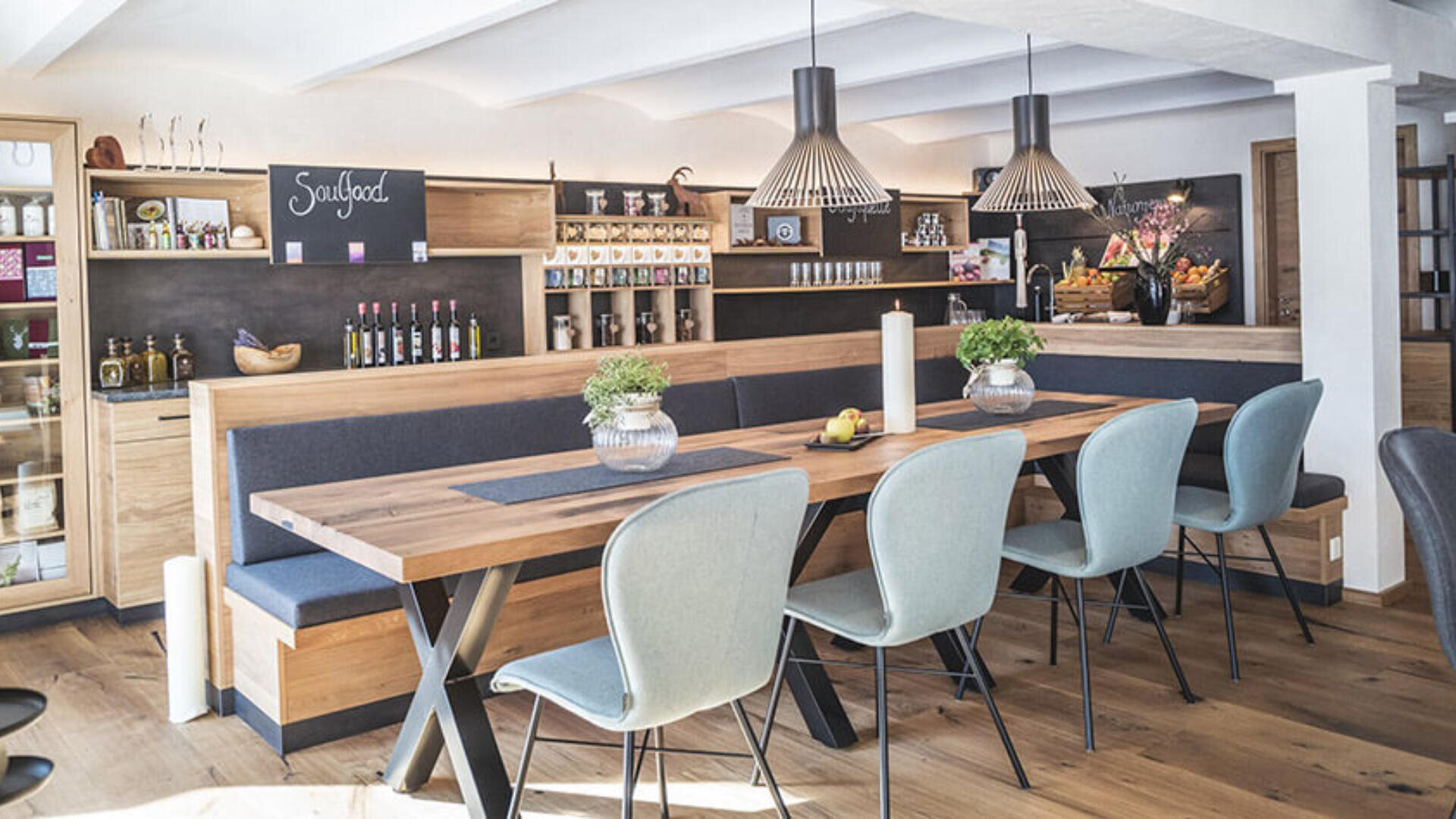 Moderner Essbereich mit langem Holztisch, blauen Stühlen, Sitzbänken und Pendelleuchten. Regale mit Flaschen, Gläsern und Deko säumen die Rückwand; „Soulfood“ steht auf einer Kreidetafel. Warme, einladende Atmosphäre.