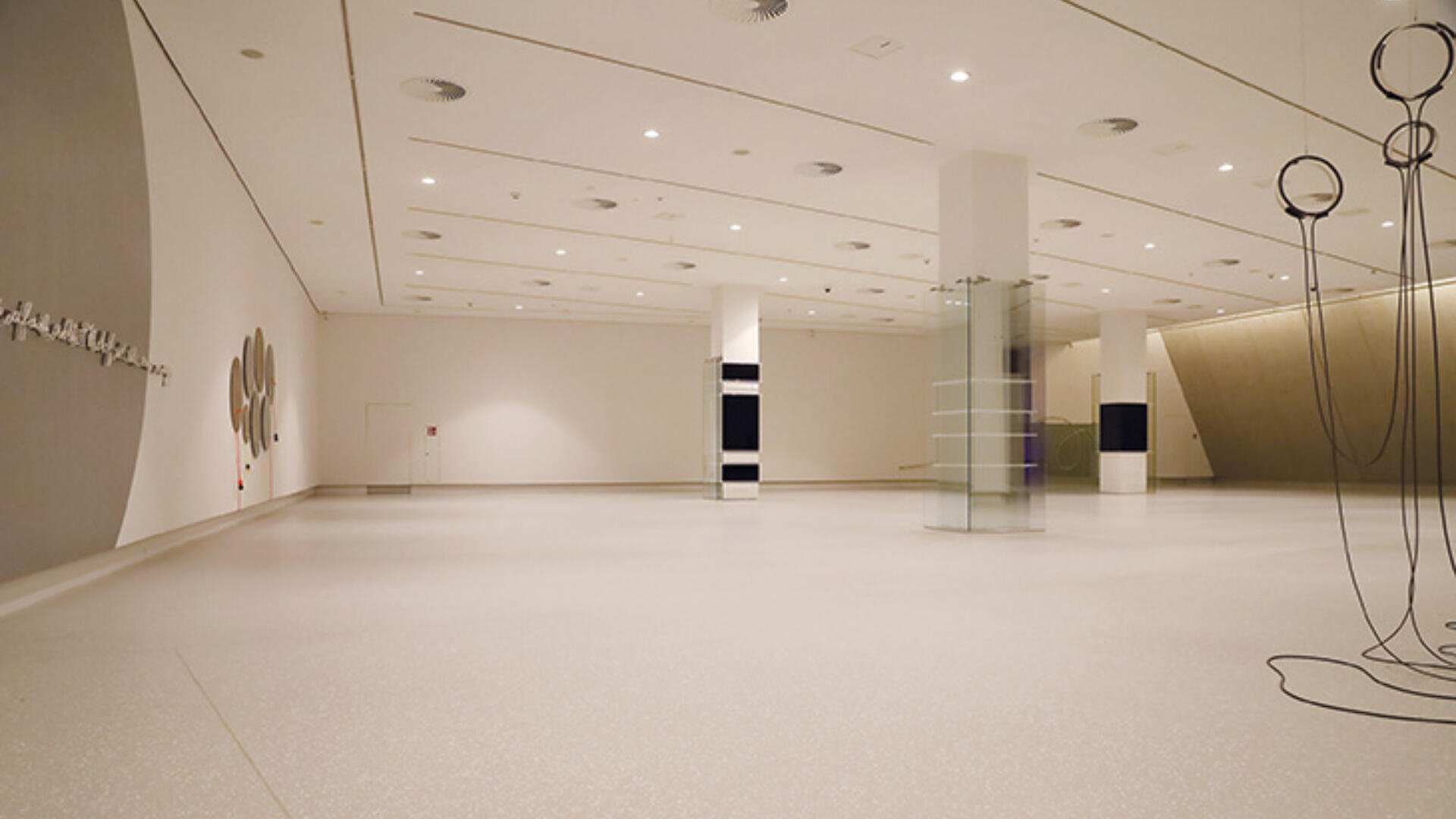 Eine geräumige, moderne Kunstgalerie mit minimalistischem Dekor, weißen Wänden, polierten hellen Böden und mehreren abstrakten Skulpturen und Wandinstallationen. Der Raum ist hell durch Deckenleuchten erleuchtet.