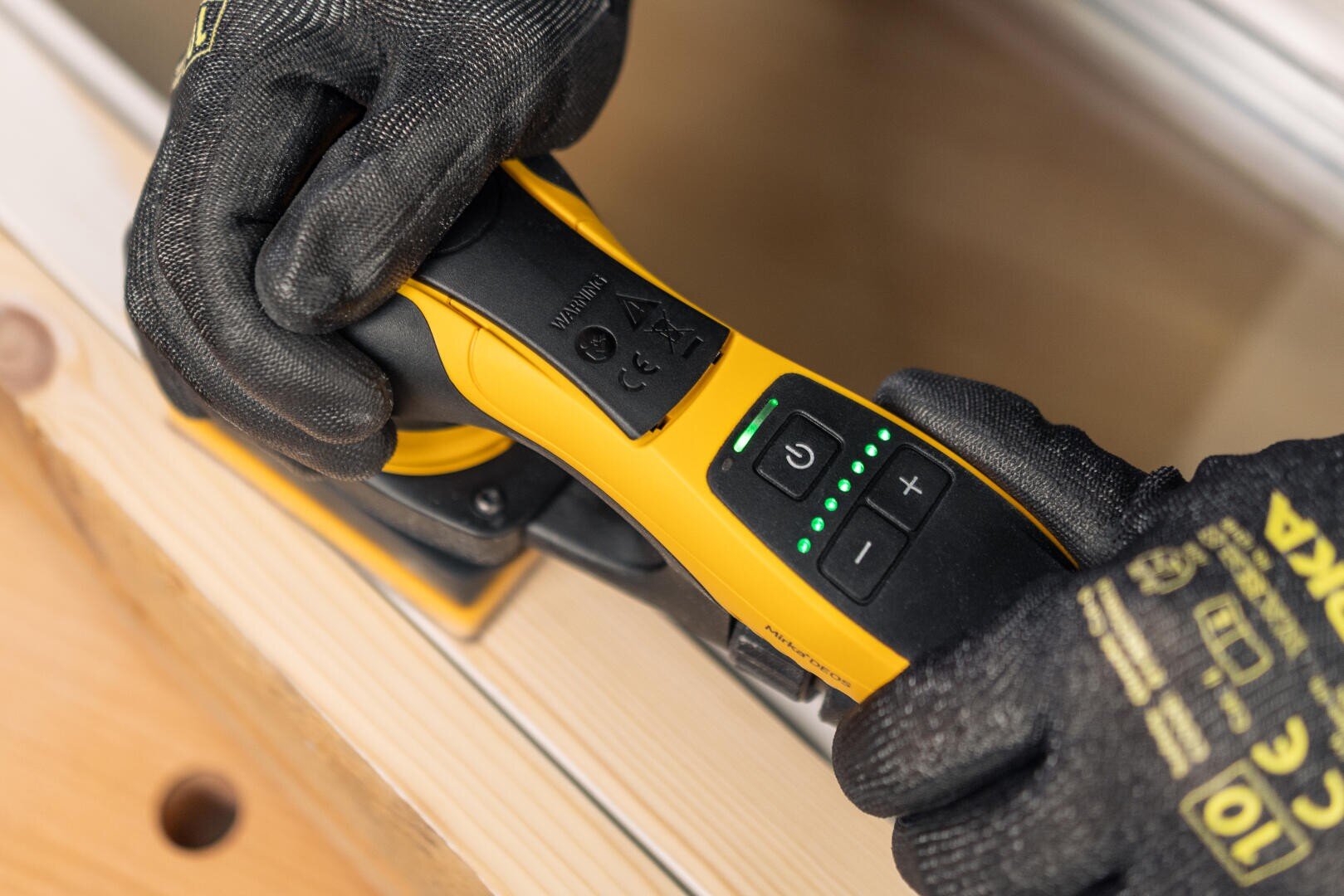 Eine Person mit schwarzen Schutzhandschuhen bedient die Bedienelemente eines gelb-schwarzen Elektrowerkzeugs, das auf einem hellen Holzstück liegt. Das Werkzeug verfügt über grüne Kontrollleuchten und Plus-/Minus-Tasten.