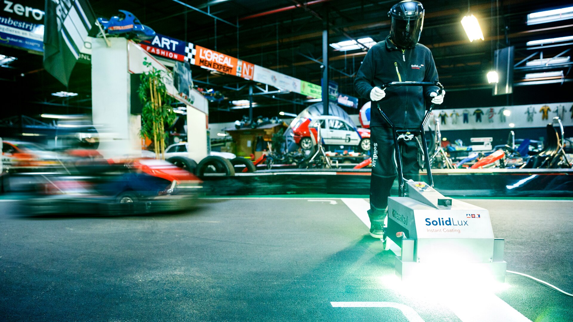 A person in protective gear operates a SolidLux machine that paints a glowing white line on an indoor go-kart track, while a go-kart speeds by in the background.