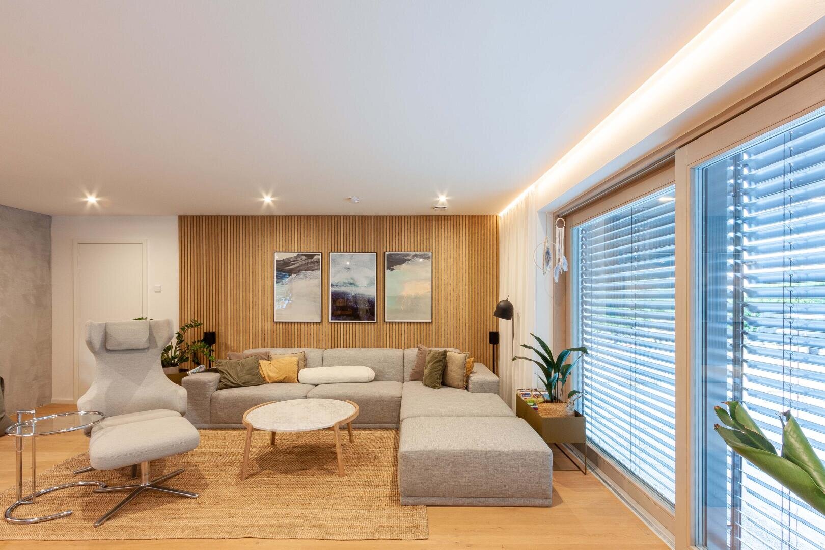 Ein modernes Wohnzimmer mit einem grauen Sofa, einem weißen Sessel, einem runden Couchtisch und drei gerahmten Kunstwerken an einer Holzwand. Große Fenster mit Jalousien lassen Tageslicht herein, und im Raum sind Pflanzen verteilt.