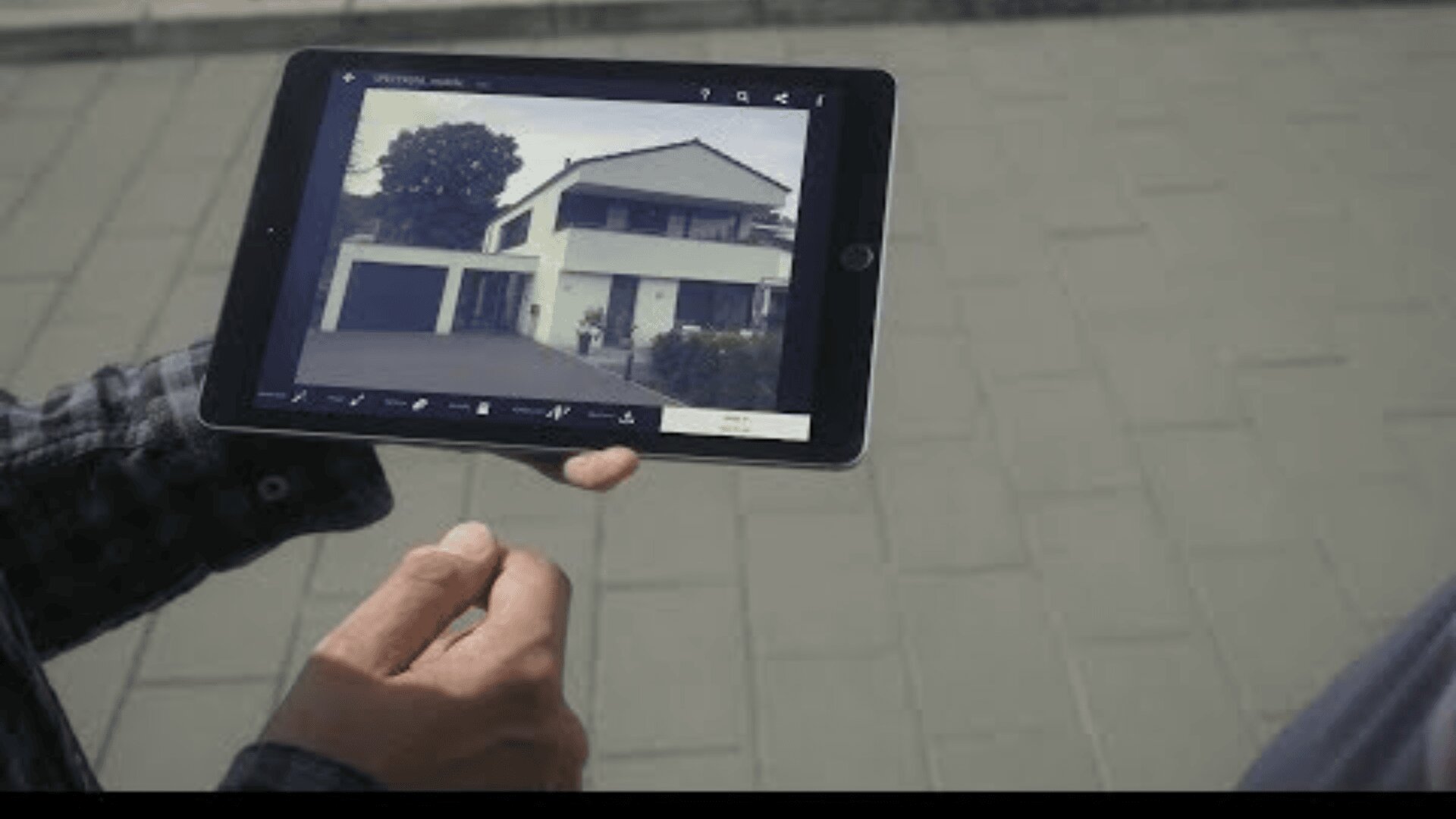 Eine Person hält ein Tablet mit dem Foto eines modernen zweistöckigen Hauses mit Garage, das im Freien auf einer gepflasterten Fläche steht.