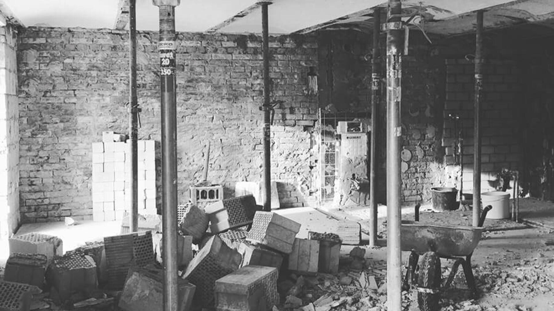 Ein Schwarzweißfoto eines im Bau befindlichen Gebäudes, auf dem Metallstützpfeiler, verstreute Ziegelsteine, Betonblöcke, Schutt auf dem Boden und eine Schubkarre neben unfertigen Ziegelwänden zu sehen sind.