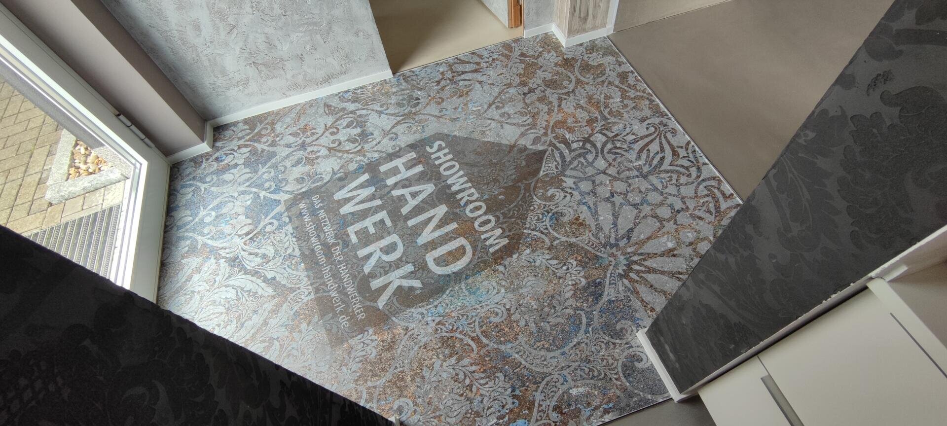 Ein dekorativer Fliesenboden im Eingangsbereich zeigt von oben den Schriftzug „SHOWROOM HANDWERK“ mit floralem Muster. Die Tür ist geöffnet, und Licht fällt ein, wodurch das aufwendige Bodendesign hervorgehoben wird.