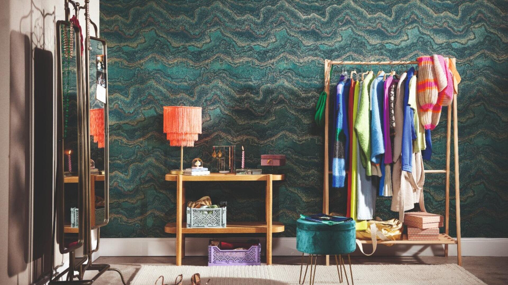 A stylish room with a green wavy-patterned wallpaper, a wooden console table with a fringed orange lamp, a standing mirror, a clothing rack with colorful clothes, and decorative boxes and a teal ottoman on the floor.