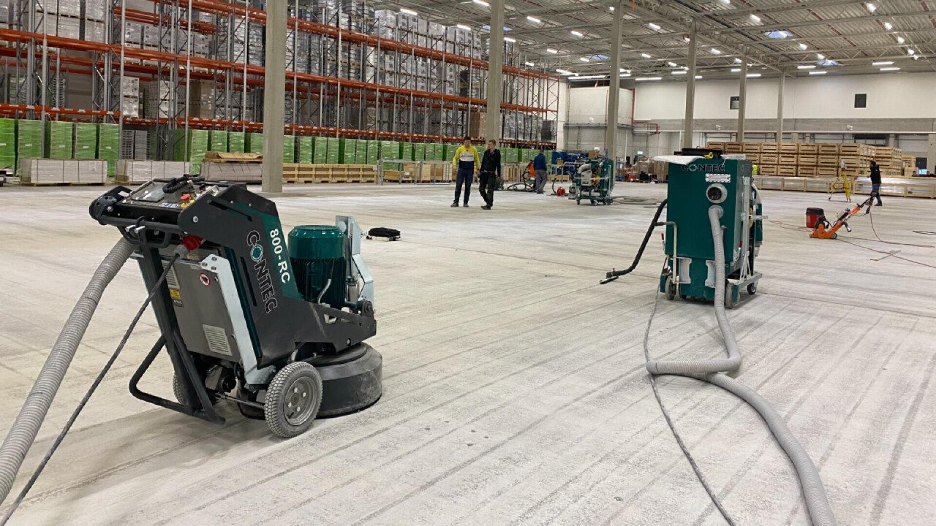 Large warehouse interior with industrial floor cleaning and polishing machines in use. Workers in safety vests are scattered throughout the space, and shelving is visible along the walls.