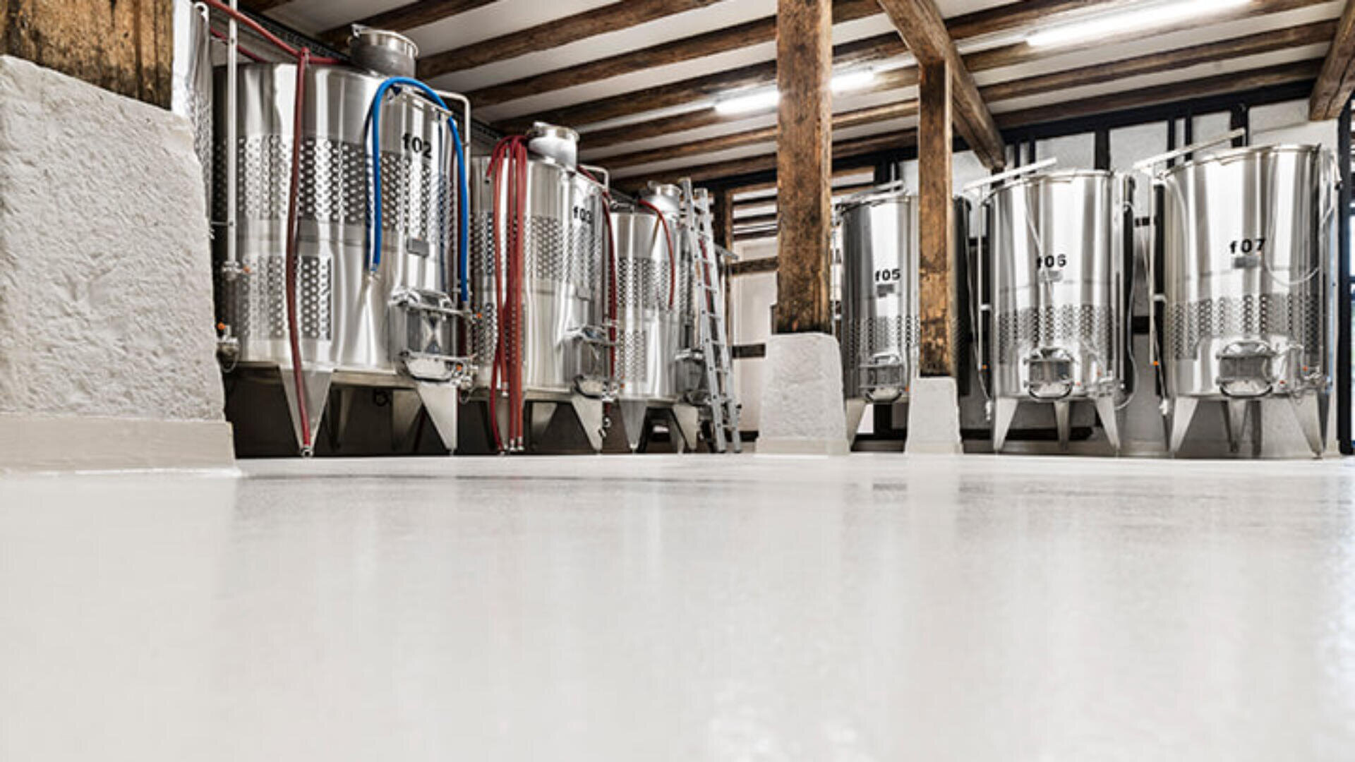 Gärtanks aus Edelstahl säumen einen sauberen, geräumigen Raum mit Holzbalken und weißen Wänden und lassen an eine moderne Brauerei oder Weinproduktionsanlage denken.