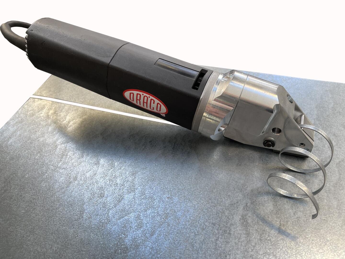 Eine schwarz-silberne elektrische Metallschere mit dem Markennamen Draco auf der Seite, die auf einem Blech liegt, neben dem Schneidkopf befinden sich aufgerollte Metallspäne.