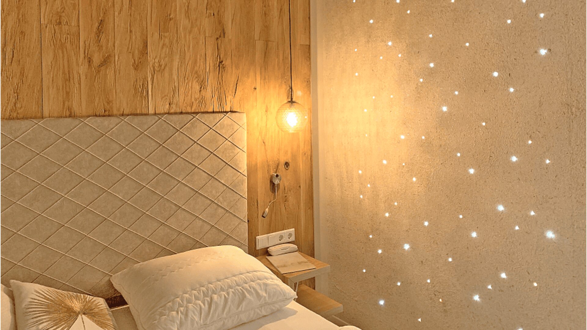 A cozy bedroom with a wooden headboard, quilted beige bed, white pillows, a hanging round lamp, and a feature wall with small glowing star-like lights.