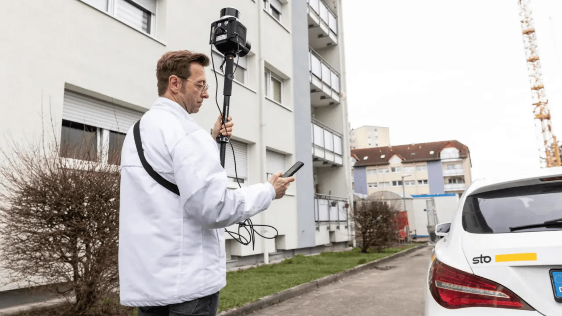 A person in a white jacket holds a pole-mounted camera and looks at a device, standing near an apartment building and a parked car on a cloudy day.