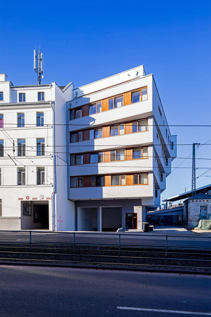 Ein modernes weißes Gebäude mit Holzfenstern steht neben einer Bahnlinie unter einem klaren blauen Himmel. Das Gebäude hat mehrere Stockwerke, Balkone und grenzt an ältere Gebäude und Bahngleise.