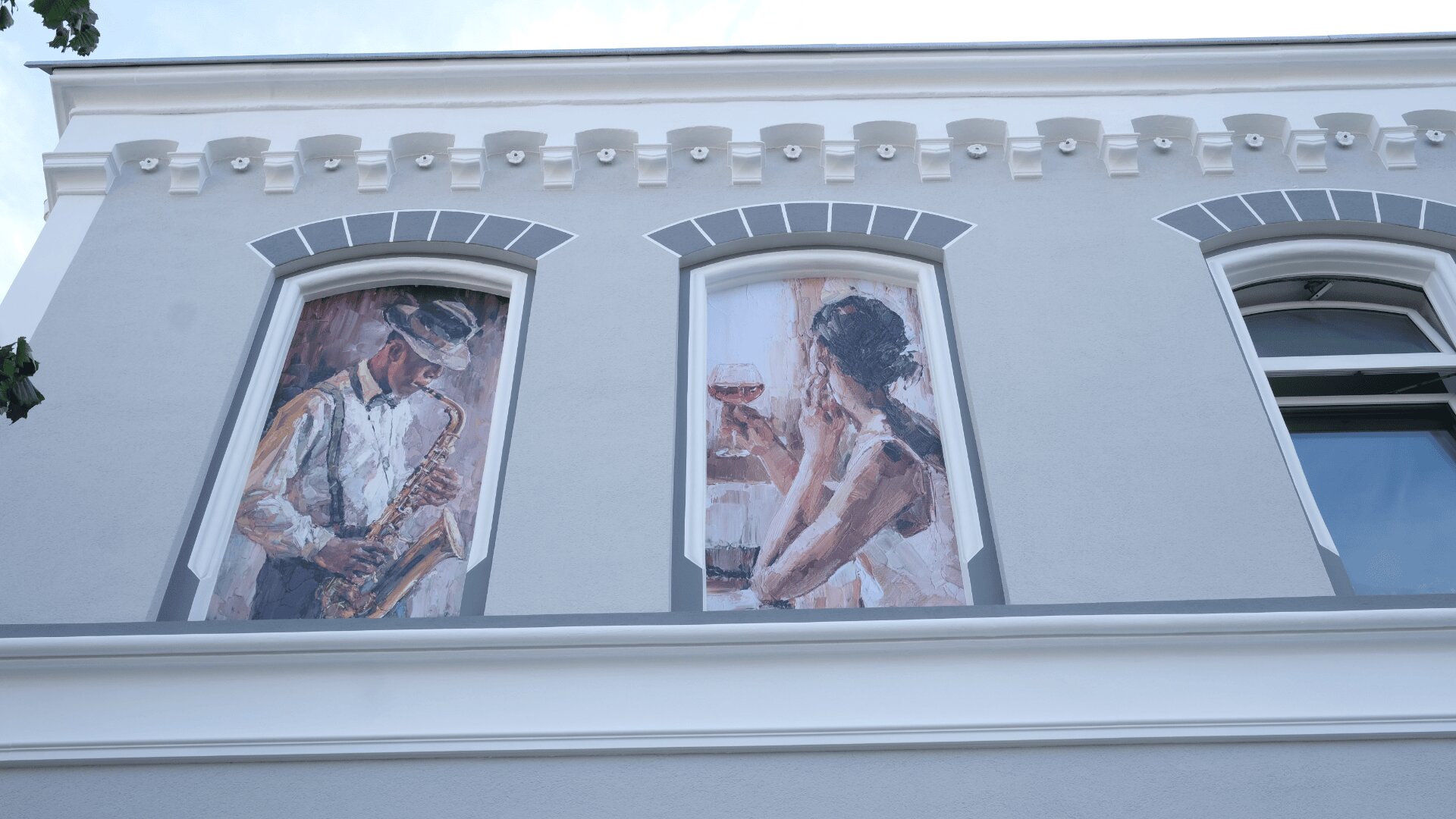 Zwei große Fenstergemälde an einem grauen Gebäude zeigen einen Saxophon spielenden Mann und eine Frau mit einem Weinglas, beide im impressionistischen Stil gemalt. Das Gebäude ist mit dekorativen weißen Zierleisten versehen.
