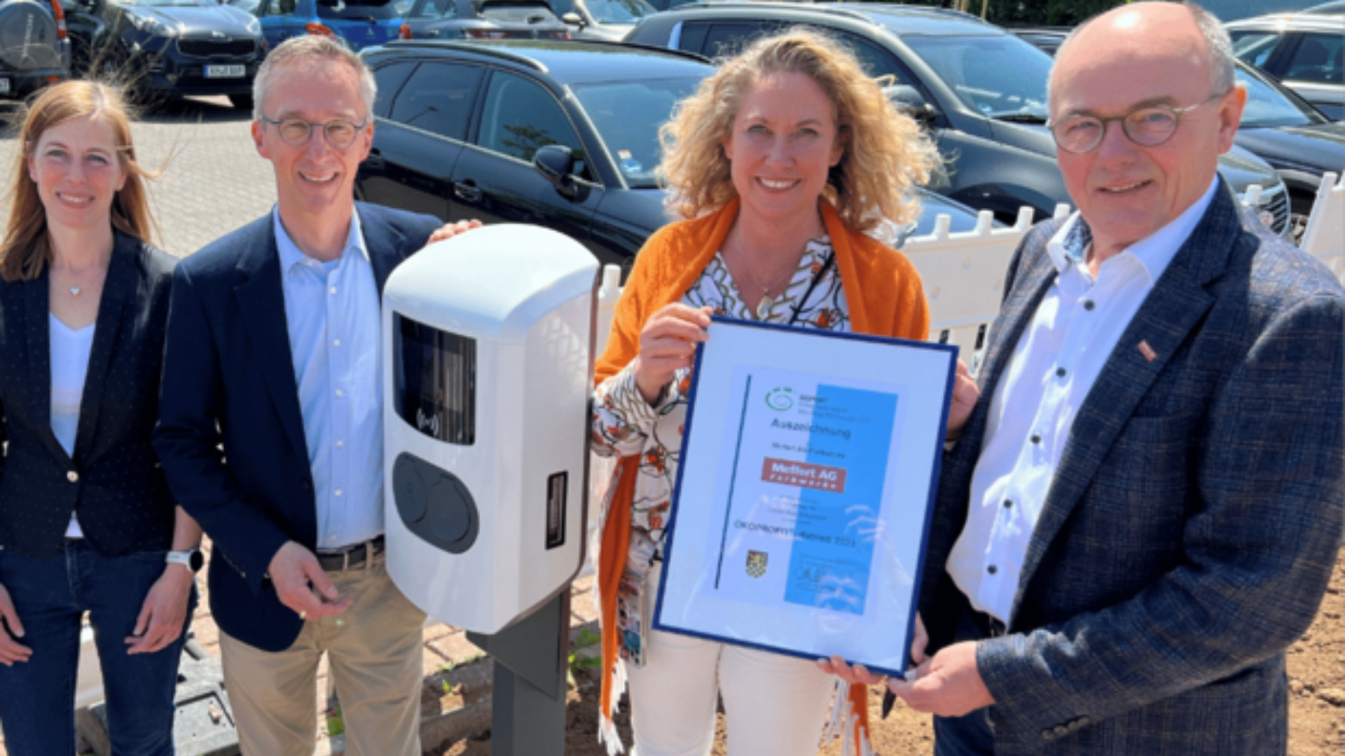 Vier Personen stehen im Freien neben einer Ladestation für Elektrofahrzeuge. Eine Frau hält eine gerahmte Urkunde in der Hand. Im Hintergrund sind Autos geparkt, und alle vier Personen lächeln in die Kamera.