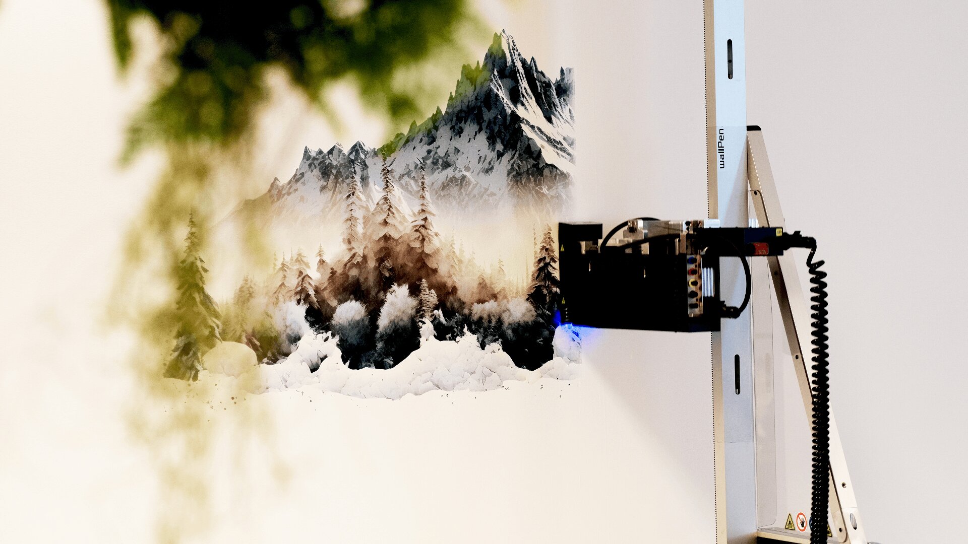 Ein Wanddrucker erstellt ein Wandbild mit schneebedeckten Bergen und Kiefern vor einer weißen Wand, wobei im Vordergrund etwas Grün verschwommen ist.
