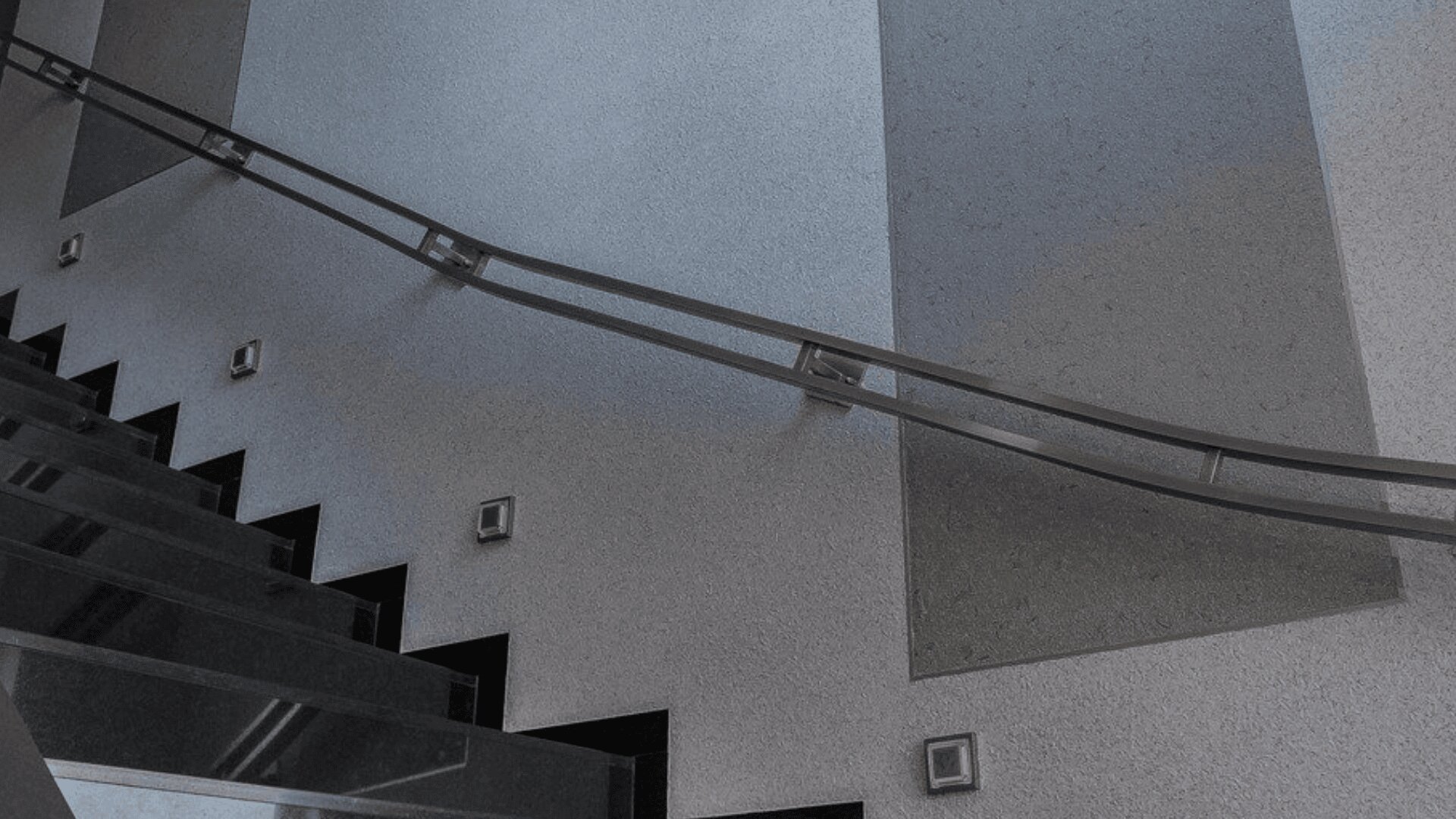 Eine moderne Treppe mit dunklen Stufen und einem glatten Metallhandlauf vor einer hellgrauen strukturierten Wand mit kleinen quadratischen Lichtern, die entlang der Wand in der Nähe der Treppe eingelassen sind.