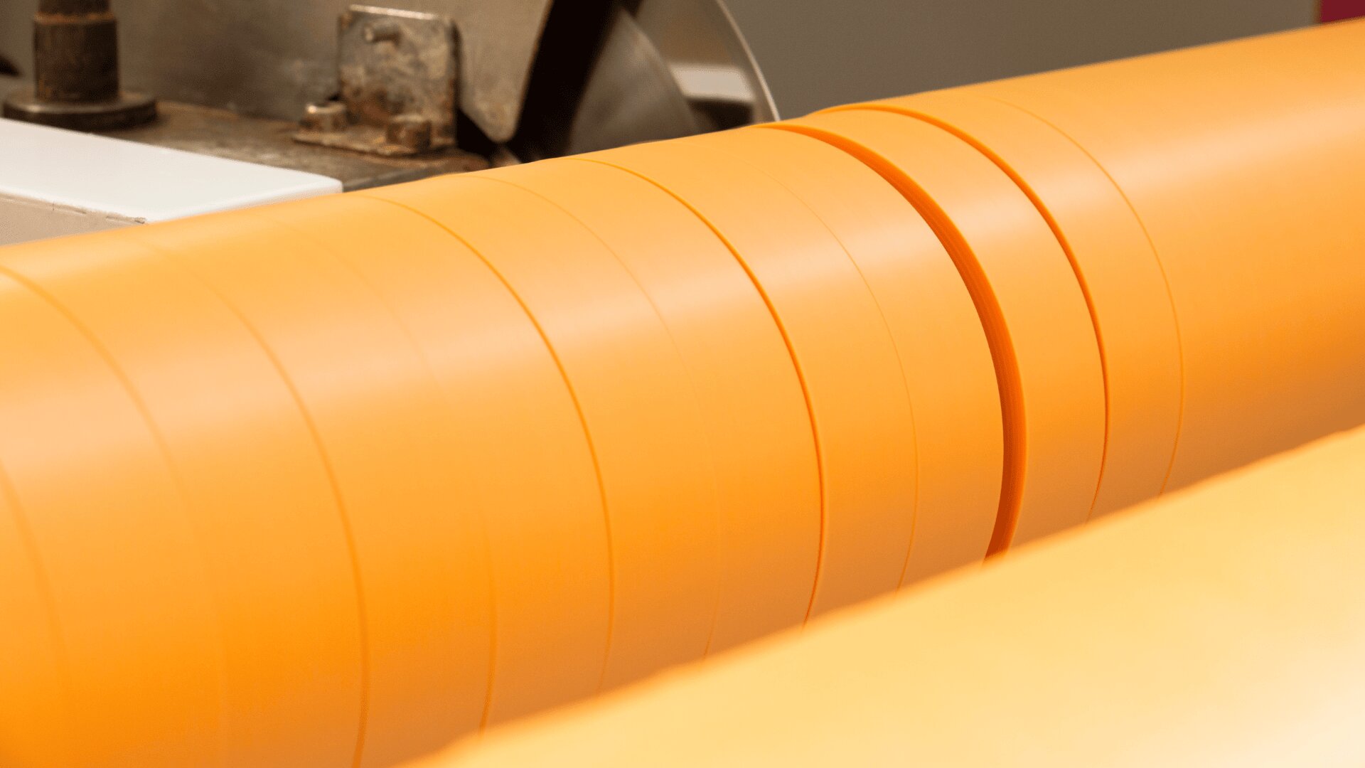 Close-up of a machine cutting or slicing a large orange plastic or rubber roll into thinner strips, showing the smooth surface and precise divisions.