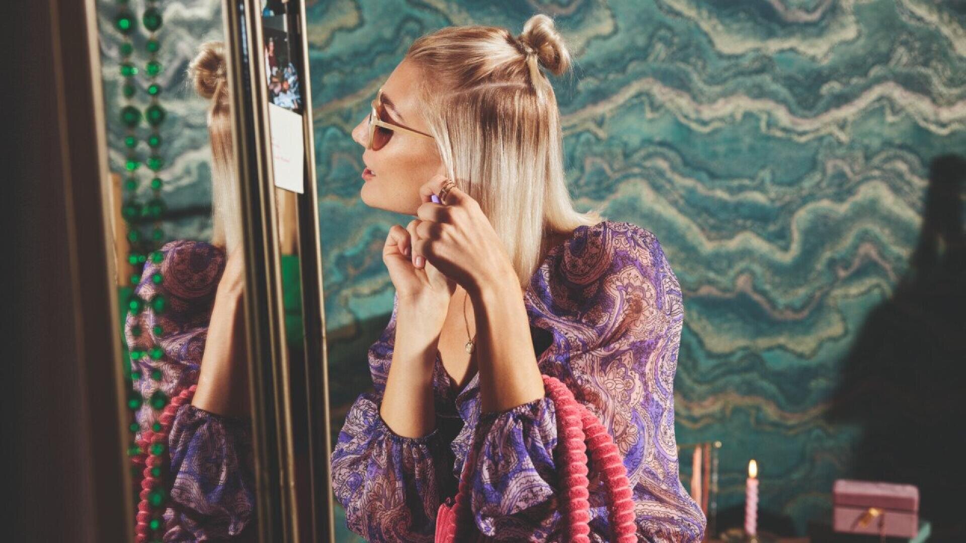 A woman with blond hair in half-up buns, wearing sunglasses and a patterned purple blouse, puts on earrings while looking in a mirror. She holds a pink textured bag; the background has a teal wavy pattern.