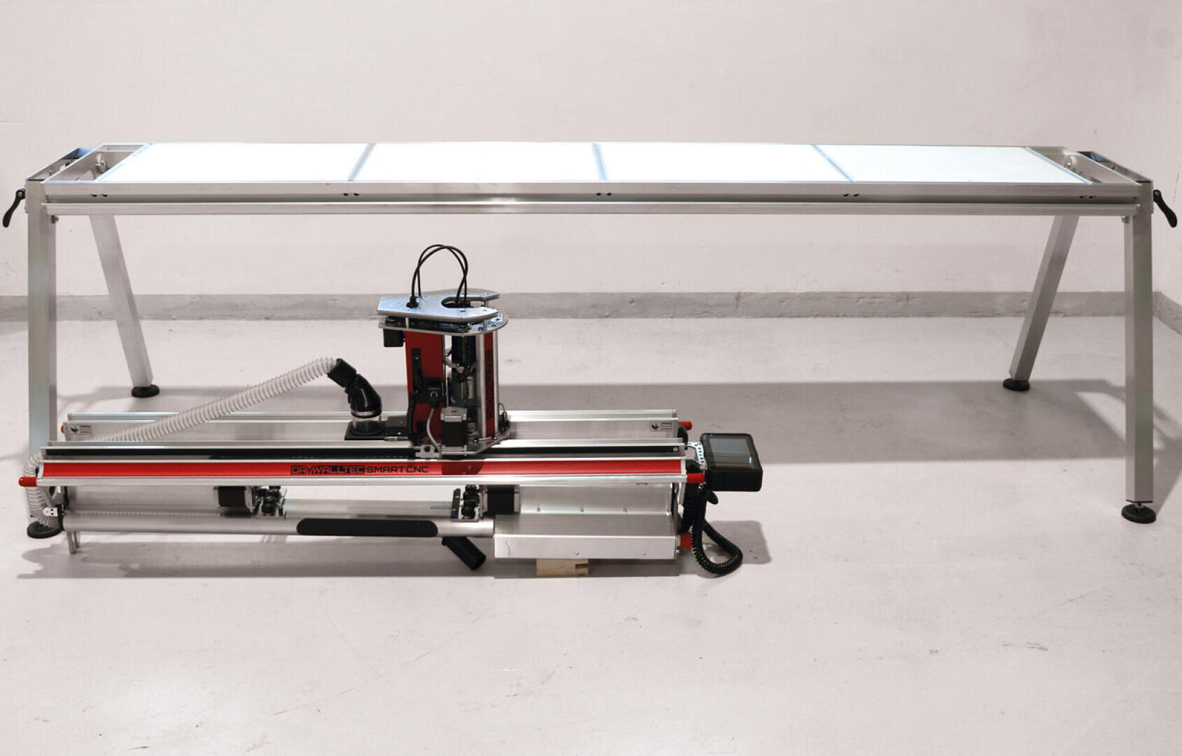 A CNC router machine with a metallic frame, red and black components, and a vacuum hose attachment is positioned in front of a long, rectangular table with a metal frame and a white tabletop.