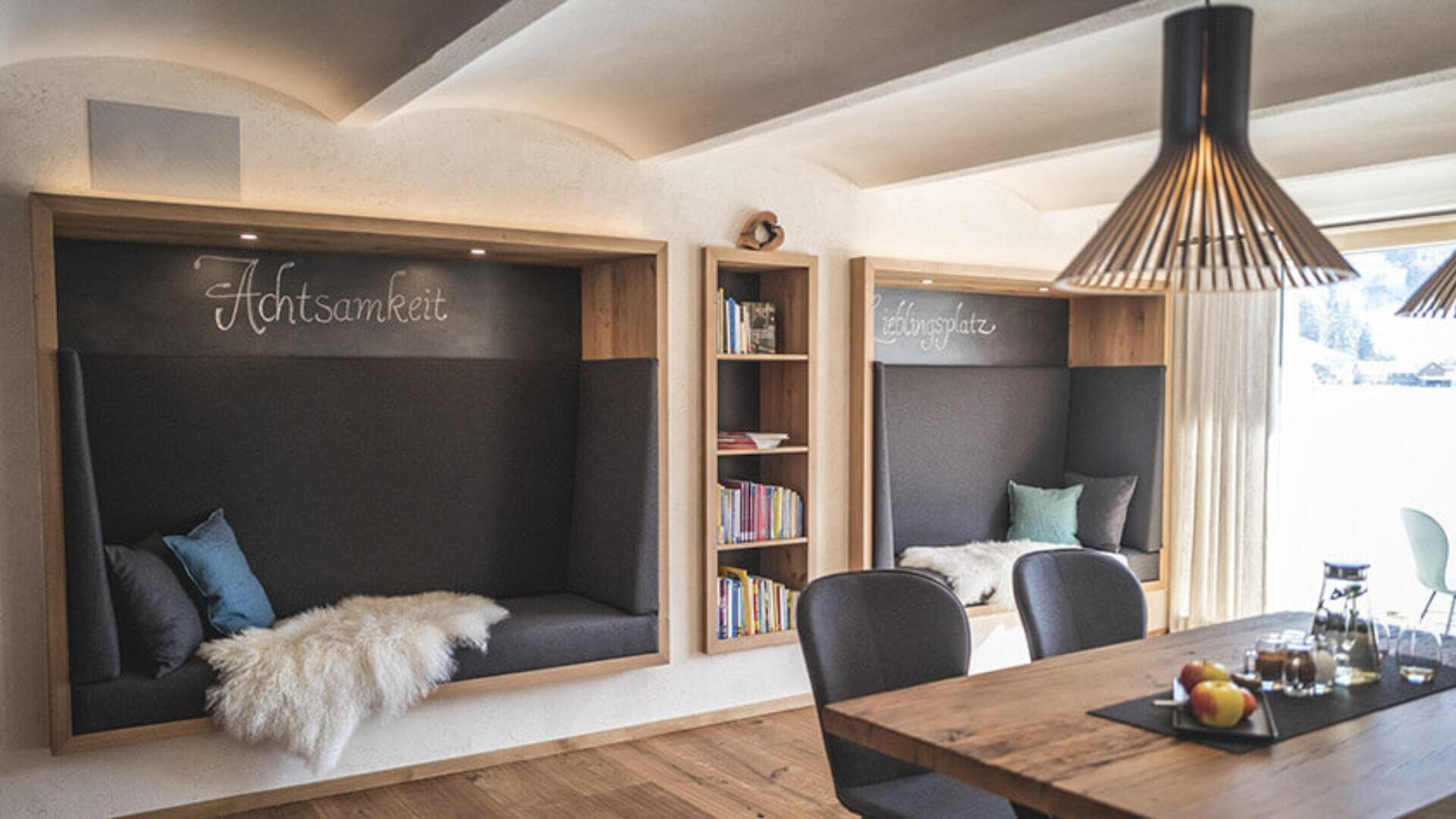 Gemütliche, moderne Leseecke mit zwei eingebauten Holzbänken, Bücherregalen, weichen Kissen und Schaffelldecken. Im Vordergrund steht ein Tisch mit Obst und Getränken unter einer stilvollen Hängelampe. Warmes, natürliches Licht durchflutet den Raum.