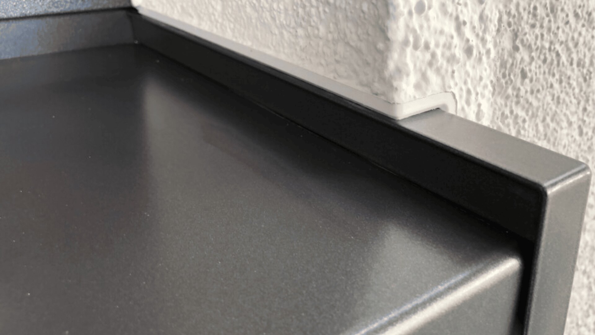Close-up of a smooth, dark gray metal surface with a raised edge, adjacent to a textured white wall. The metal edge has a notch that fits around a protrusion in the wall.