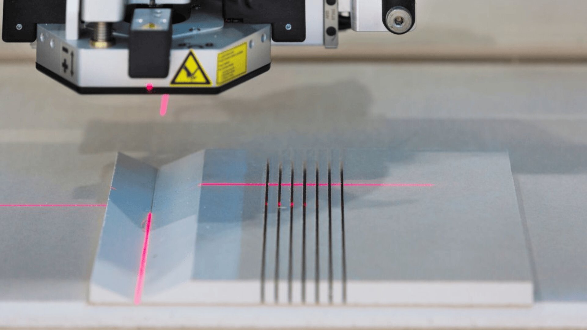 Eine Maschine mit einem Laser und einem Warnschild schneidet gerade Linien in ein flaches graues Material, wobei auf der Oberfläche rote Laserlinien zu sehen sind.
