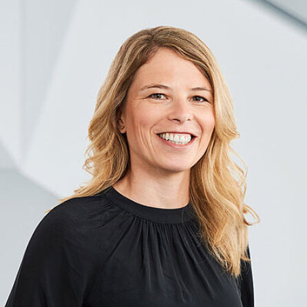 Eine Frau mit langen blonden Haaren und einem schwarzen Top lächelt in die Kamera und steht vor einem hellen, unscharfen Hintergrund.