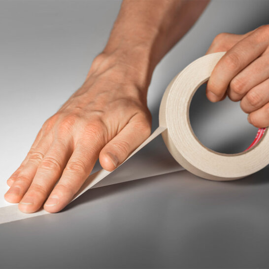 Die Hände einer Person ziehen einen Streifen Klebeband von einer Rolle ab und drücken ihn auf eine glatte, graue Oberfläche.
