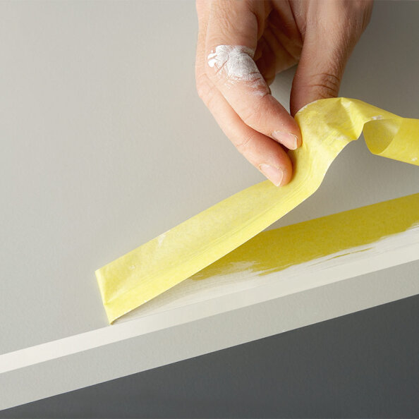 Eine Hand mit weißer Farbe an den Fingern zieht gelbes Malerklebeband vom Rand einer weißen Oberfläche ab und gibt eine saubere Farblinie frei.