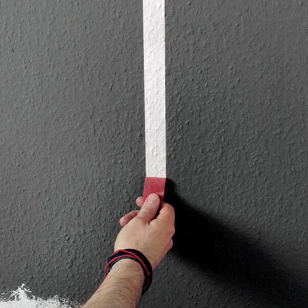 Eine Hand zieht rotes Malerband von einer grauen strukturierten Wand ab und gibt unter dem Band eine gerade, weiß gemalte Linie frei.